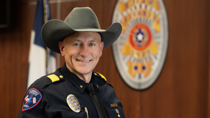 East Texas alum Arley Sansom, dressed in his police uniform and wearing his new chief's badge, smiles for a professional photo against a woodgrain wall featuring a large, out-of-focus Terrell Police Department seal.